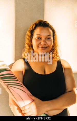 Porträt einer üppigen Frau, die eine Trainingsmatte an die Wand hält Stockfoto