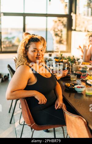 Porträt einer lächelnden Frau in Übergröße, die auf einem Stuhl sitzt und am Esstisch frühstückt Stockfoto