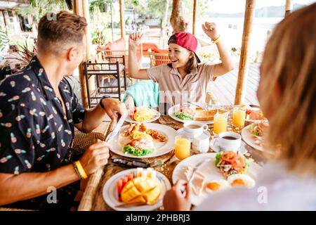 Ein glückliches Mädchen, das die Fäuste knallte, während die Eltern am Tisch im Resort frühstückten Stockfoto