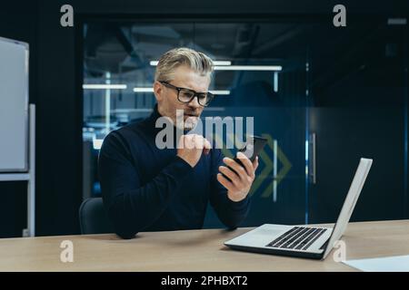 Ein gravierender, grauhaariger Geschäftsmann, der im Büro am Schreibtisch sitzt und ein Handy benutzt. Liest Nachrichten, schreibt Nachrichten, überprüft Post. Stockfoto