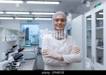 Portrait einer jungen muslimischen Labortechnikerin, Apotheker in Hijab und Schutzbrille, im medizinischen Labor, Arme gekreuzt auf der Brust, lächelnd vor der Kamera. Stockfoto