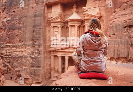 Junge Frau in Winterjacke sitzt auf einem kleinen roten Kissen am Aussichtspunkt und blickt auf Al-Khaznov - Treausry, Hauptattraktion von Petra, Jordanien. Blick von Behin Stockfoto
