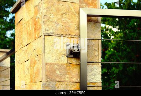 Zaunpfahl aus Naturstein. pier mit Edelstahltor und Scharnier. Geschweißter Vierkantrohrrahmen. Horizontale verdrillte Kabel. Grün. Strauchhintergrund Stockfoto