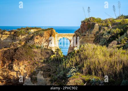 Abendlicher Blick auf die Ponte Romana de Lagos, eine bogenförmige Steinbrücke, die zwei Klippen mit dem Horizont des Atlantischen Ozeans im Hintergrund verbindet Stockfoto