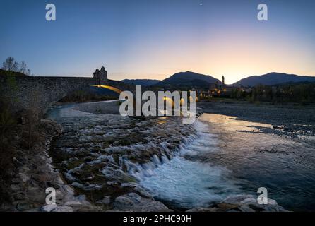 Nächtliche Luftaufnahme der alten mittelalterlichen Brücke, genannt „Ponte del Diavolo“ oder „Ponte Gobbo“, auch „Teufelsbrücke“ über dem Fluss Trebbia nach Sonnenuntergang, Stockfoto