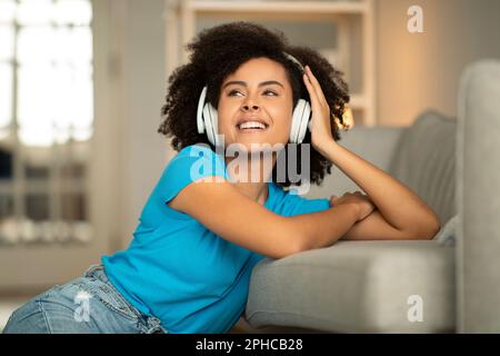 Die nette junge, lockige afroamerikanerin mit kabellosen Kopfhörern hört Musik und genießt die Freizeit Stockfoto