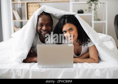 Fröhlicher afroamerikanischer Mann und Frau, die unter einer Leinendecke mit einem Laptop auf dem Bett in einem schönen Apartment wohnten. Glückliche Familie, die Spaß zusammen hat und soziale Medien auf einem modernen Gerät überprüft. Stockfoto