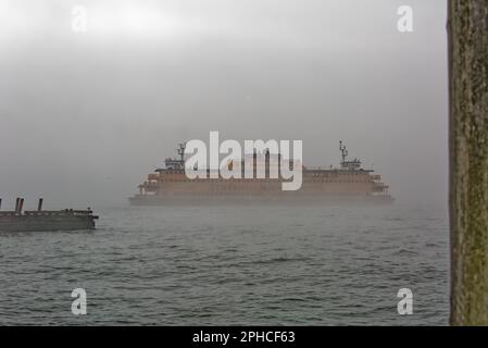 Die Staten Island Ferry ist der einzige Farbfleck an einem nebligen Februar Morgen im Hafen von New York. Der Senator John J. Marchi verlässt Manhattan. Stockfoto