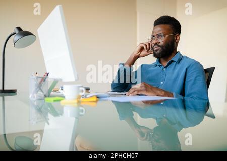 Konzentrierter Schwarzer, der im Büro an einem Computer arbeitet, Projekte überprüft, alltägliche Probleme löst oder ein Video-Meeting abhält Stockfoto