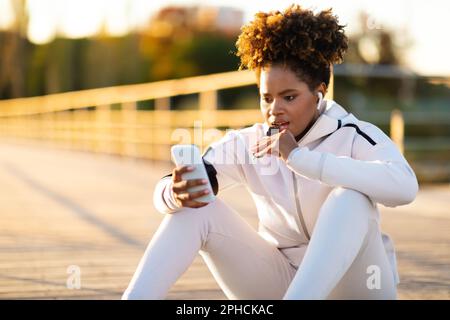 Junge, Sportliche Schwarze Frau, Die Proteinriegel Isst Und Smartphone Im Freien Benutzt Stockfoto