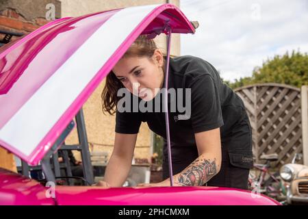 Junge Mechanikerin, die Wartungsarbeiten an einem leuchtend rosa Klassiker Austin Mini durchführt Stockfoto