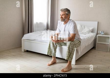 Positiv ausgeruhter reifer Mann, der auf dem Bett sitzt und Tee trinkt Stockfoto