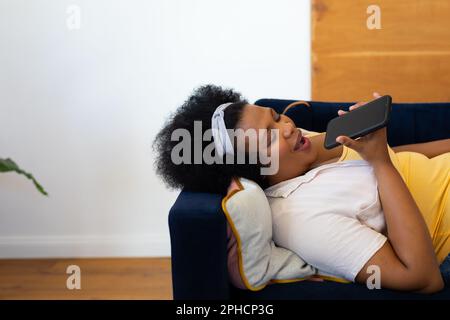 Eine glückliche afroamerikanische Frau in Übergröße, die auf dem Sofa liegt und mit dem Smartphone spricht Stockfoto