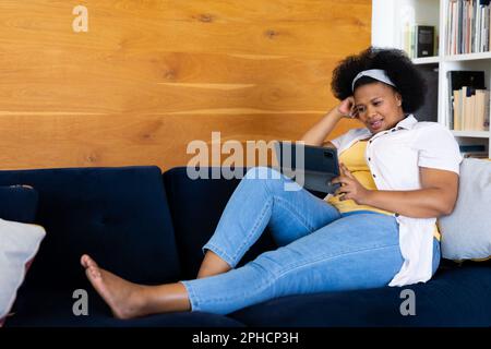 Eine glückliche afroamerikanische Frau in Übergröße, die sich auf dem Sofa entspannt und ein Tablet benutzt Stockfoto