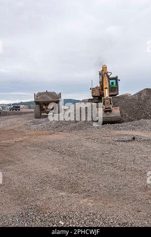 Ein Bagger zieht Steine und Schmutz in der Nähe eines Kipplasters auf einer aktiven Deponie hoch. Stockfoto