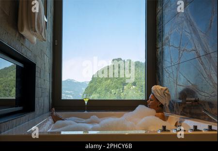 Frau liegt in einer Badewanne mit einem Glas Champagner und genießt den Fensterblick über den Berg an einem sonnigen Sommertag in Burgenstock, Nidwalden, Switzerlan Stockfoto