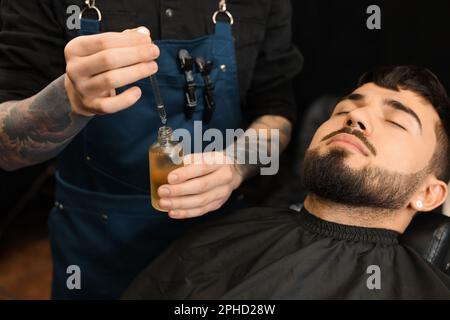 Friseur mit Bartöl in der Nähe des Kunden im Friseursalon, Schließung. Professioneller Rasierservice Stockfoto