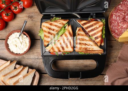 Moderne Grillmaschine mit Sandwiches und verschiedenen Produkten auf Holztisch, flach liegend Stockfoto