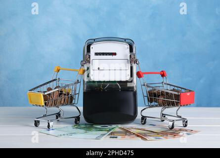 Stromzähler, kleine Einkaufswagen mit Münzen und Euro-Banknoten auf weißem Holztisch Stockfoto