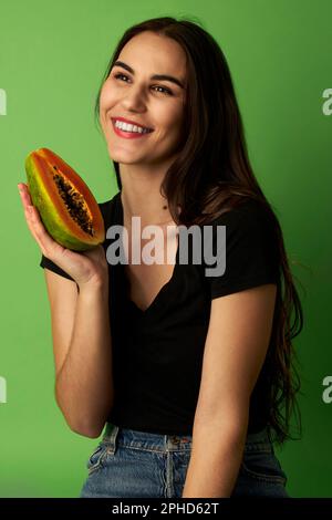 Eine Frau, die eine geschnittene Papaya in einem schwarzen Hemd hält, vor einem grünen Hintergrund steht und auf die Nahaufnahme schaut Stockfoto