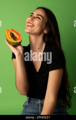 Eine Frau, die eine geschnittene Papaya hält und ein schwarzes Hemd trägt. Sie steht vor einem grünen Hintergrund und schaut nach oben Stockfoto