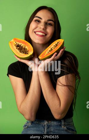 Eine Frau, die eine geschnittene Papaya in einem schwarzen Hemd hält, vor einem grünen Hintergrund steht und vor der Kamera lächelt Stockfoto