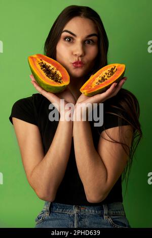 Eine Frau, die eine geschnittene Papaya in einem schwarzen Hemd hält, vor einem grünen Hintergrund steht und in die Kamera schaut Stockfoto