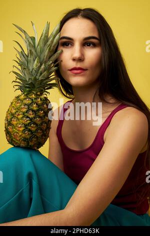 Eine Frau, die sich eine Ananas auf den Kopf lehnt und ein rotes Hemd vor einem gelben Hintergrund trägt Stockfoto