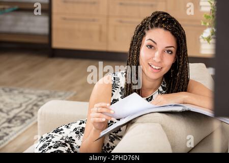 Nahaufnahmen einer jungen Frau, die lächelt und fasziniert und leicht überrascht ist, während sie durch ein Notizbuch oder einen Katalog blättert und sich gegen ihr Zuhause lehnt Stockfoto