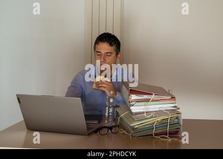 Bild eines verzweifelten Mannes mit einem Sandwich und einer kleinen Flasche Wasser, der wegen Überstunden in seinem Büro arbeiten muss. Stockfoto