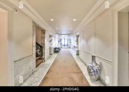 Ein langer Flur mit weißen Wänden und beigefarbenem Teppich auf dem Boden, der zu einer offenen Tür führt, die zu einem anderen Zimmer führt Stockfoto