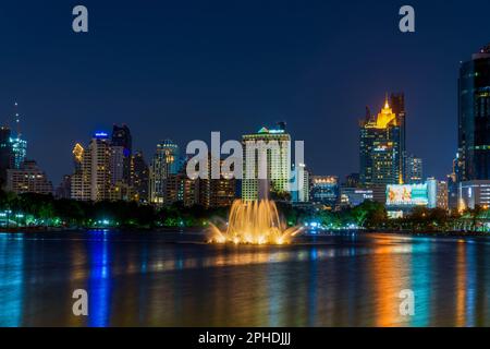 Bangkoks Skyline in einer klaren Nacht vom Benchakitti Park aus gesehen - im Vordergrund befindet sich der ruhige See und der glühende Brunnen des Parks. Stockfoto