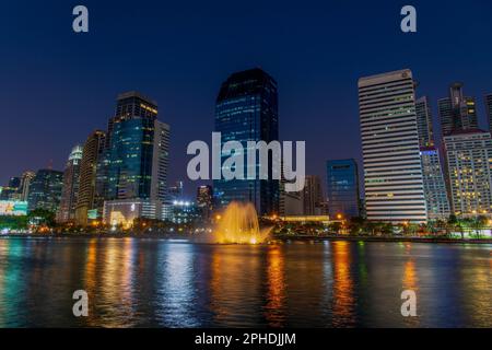 Bangkoks Skyline in einer klaren Nacht vom Benchakitti Park aus gesehen - im Vordergrund befindet sich der ruhige See und der glühende Brunnen des Parks. Stockfoto