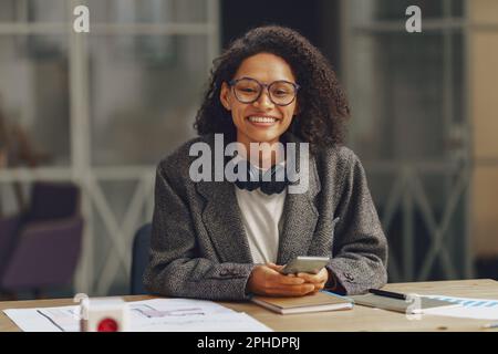 Freiberuflerin mit Kopfhörern, die vor der Kamera lächelt, während sie in moderner Zusammenarbeit mit dem Telefon sitzt Stockfoto