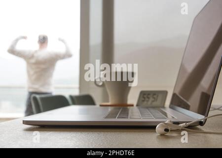 Ein digitaler Nomadenmann beendete die Arbeit und feierte auf dem Balkon. Arbeitsplatz mit Kaffeetasse, Laptop und Kopfhörern Stockfoto