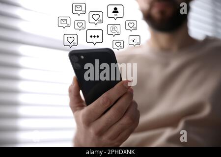 Verschiedene virtuelle Ikonen und junger Mann, der sein Smartphone drinnen benutzt, Nahaufnahme. SMM-Konzept (Social Media Marketing) Stockfoto