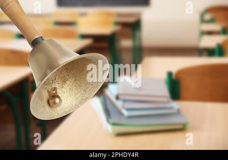 Goldene Schulglocke mit Holzgriff und verschwommenem Blick auf Bücher auf dem Schreibtisch im Klassenzimmer Stockfoto