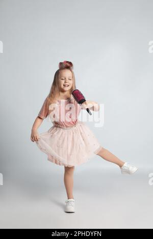 Süßes kleines Mädchen mit einer Haarbürste, die auf hellgrauem Hintergrund singt Stockfoto