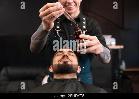 Friseur mit Bartöl in der Nähe des Kunden im Friseursalon, Schließung. Professioneller Rasierservice Stockfoto