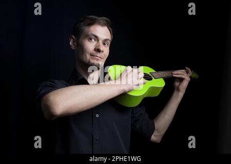 Hübsches, junger Mann-Porträt mit lustiger grüner Ukulele-Gitarre auf schwarzem Hintergrund Stockfoto