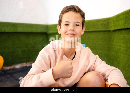 Portrait eines attraktiven, fröhlichen, lächelnden 11-jährigen Jungen, draußen über einer grünen Wand. Glücklicher, gutaussehender Teenager mit Daumen nach oben. Süßes Kinderporträt. Stockfoto