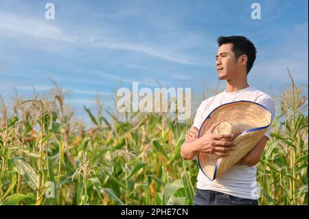 Erfolgreicher männlicher Millenialer asiatischer Bauer oder Besitzer einer Maisfarm mit Strohhut in der Hand steht mit gekreuzten Armen auf seinem Maisfeld. Stockfoto