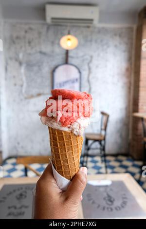 Eiskrem oder Eis in der linken Hand. Von rechts fotografiert. Mit einem Hintergrund aus Cafétischen und Stühlen. Stockfoto