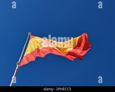 Spanische Nationalflagge auf blauem Himmelshintergrund. Die Fahne am Fahnenmast flattert im Wind. Spanische Flagge bläst im Wind gegen klares Blau Stockfoto