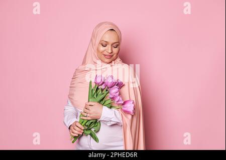 Selbstbewusste, schwangere, muslimische Frau in rosa Hijab, lächelt mit einem Strauß lila Tulpen auf isoliertem rosa Hintergrund Stockfoto