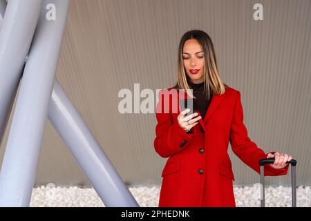 Junge glückliche Frau in dunkelblondem Haar und rotem Lippenstift, die einen roten Mantel trägt und mit dem Koffer steht, während sie in der Nähe grauer Stangen auf dem Handy surft Stockfoto