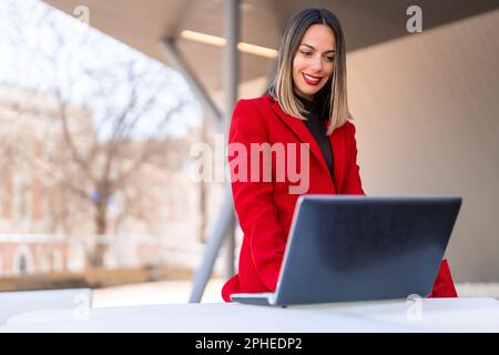 Glückliche junge Frau mit dunkelblondem Haar und rotem Lippenstift, die am Tisch sitzt und vor verschwommenem Hintergrund auf den Laptop-Bildschirm schaut Stockfoto