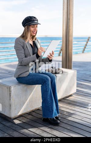 Der ganze Körper einer lächelnden jungen Frau in einer grauen Jacke, die auf einem Tablet zeichnet, während sie auf dem Seeweg im Hafen sitzt Stockfoto