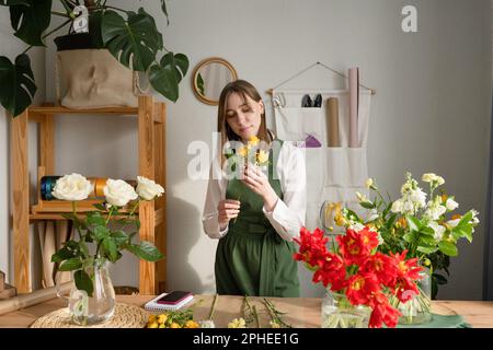 Weibliche Floristin in grüner Schürze steht am Tisch und macht während des Arbeitstages im Blumengeschäft einen Strauß frischer Blumen Stockfoto