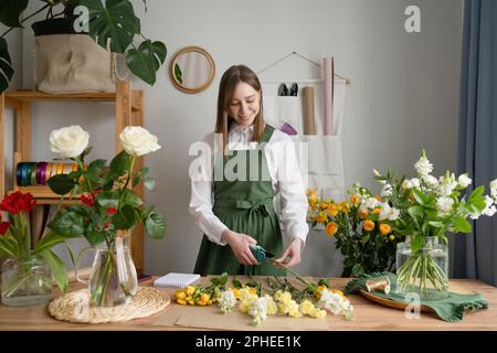 Positive junge Blumenmeisterin in grüner Schürze, die am Tisch steht und Blumen im Blumenladen schneidet Stockfoto